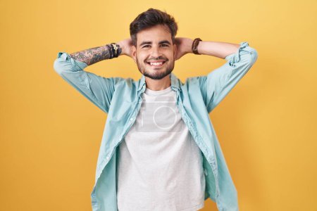 Foto de Joven hombre hispano con tatuajes de pie sobre fondo amarillo relajante y estiramiento, brazos y manos detrás de la cabeza y el cuello sonriendo feliz - Imagen libre de derechos