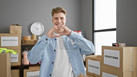 Photo for Handsome young caucasian man working hard, confidently volunteering at the heart of community charity center, his smile is infectious as he stands enjoying altruism, forming a lovely heart gesture - Royalty Free Image