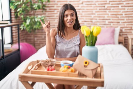 Photo for Brunette young woman eating breakfast sitting on the bed smiling happy pointing with hand and finger to the side - Royalty Free Image