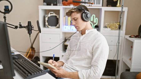 Photo for Focused young caucasian man, a handsome musician, engrossed in composing an acoustic song at a music studio, listening to melodies, notebook in hand, indoor ambiance - Royalty Free Image