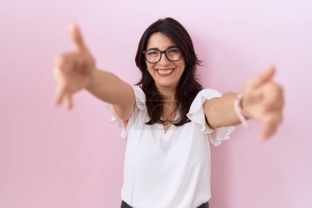 Foto de Mujer hispana de mediana edad con camiseta blanca casual y gafas mirando a la cámara sonriendo con los brazos abiertos para abrazarse. expresión alegre abrazando la felicidad. - Imagen libre de derechos