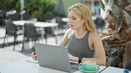 Photo for Young blonde woman having video call taking notes at coffee shop terrace - Royalty Free Image