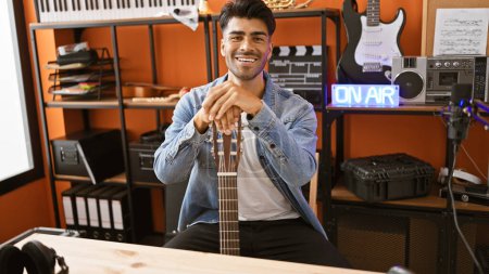 Photo for A smiling young hispanic man with a beard in a music studio, holding a guitar, sitting in front of a microphone and 'on air' sign. - Royalty Free Image
