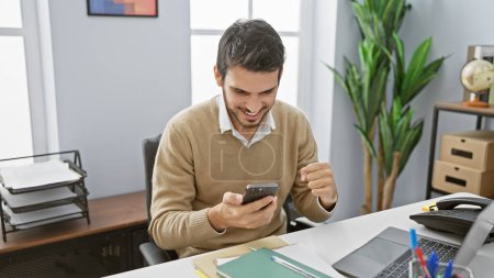 Photo for Handsome young hispanic man excitedly looks at smartphone in a modern office setting - Royalty Free Image
