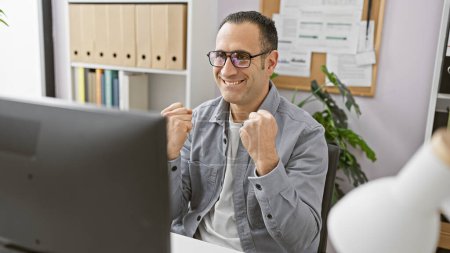 Foto de Un hombre hispano sonriente con gafas se siente logrado en su oficina organizada con una computadora y una planta. - Imagen libre de derechos