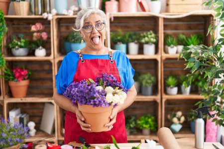 Foto de Mujer de mediana edad con pelo gris trabajando en floristería sosteniendo la planta sacando la lengua feliz con expresión divertida. - Imagen libre de derechos