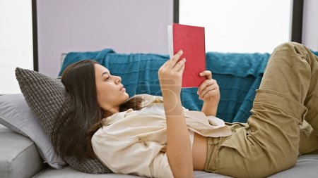 Foto de Hermosa mujer hispana joven cómodamente absorta en la lectura de un libro, acostada en el sofá en casa, que representa un retrato de relajación y ocio inteligente en el interior de su sala de estar - Imagen libre de derechos