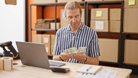 Photo for A mature businessman counts money at his warehouse workstation, looking focused and professional. - Royalty Free Image