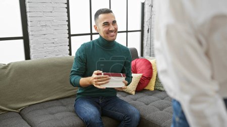 Téléchargez les photos : Un homme souriant présente un cadeau au partenaire dans un salon confortable et moderne, transmettant amour et célébration - en image libre de droit