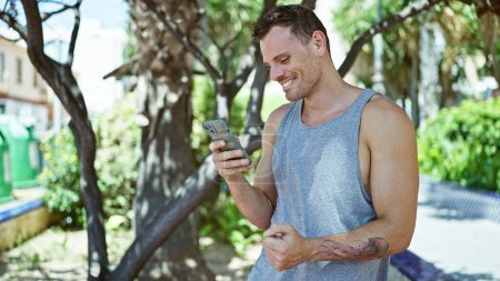 Téléchargez les photos : Bel homme hispanique souriant tout en utilisant smartphone à l'extérieur dans un parc verdoyant luxuriant - en image libre de droit