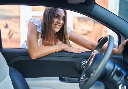 Foto de Joven hermosa mujer hispana sonriendo confiada apoyada en la puerta del auto en la calle - Imagen libre de derechos