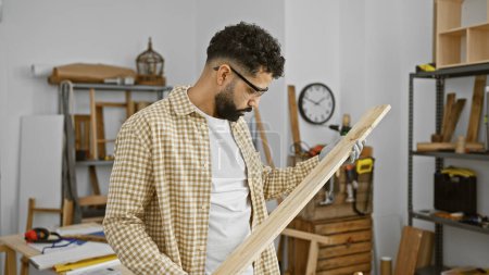 Téléchargez les photos : Bel homme hispanique avec barbe examinant le bois dans un atelier de menuiserie. - en image libre de droit