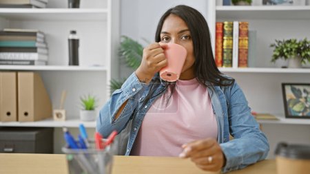 Young, pregnant business woman, blissfully sipping coffee at her office desk, embodying success while expecting motherhood.