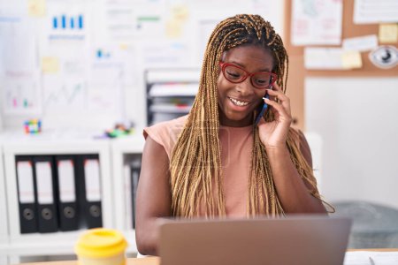 Téléchargez les photos : Femme afro-américaine travailleur d'affaires en utilisant un ordinateur portable parlant sur smartphone au bureau - en image libre de droit