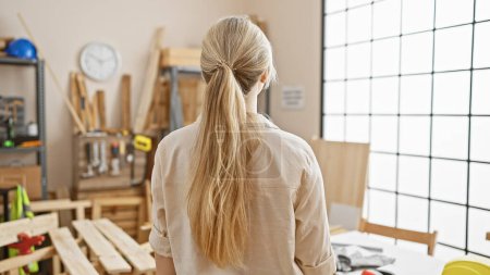 Téléchargez les photos : Vue arrière d'une jeune femme caucasienne aux cheveux blonds dans un atelier de menuiserie regardant outils et menuiserie. - en image libre de droit