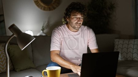 Photo for A focused young man using a laptop in a cozy living room at night, with a decorative lamp and cushion. - Royalty Free Image