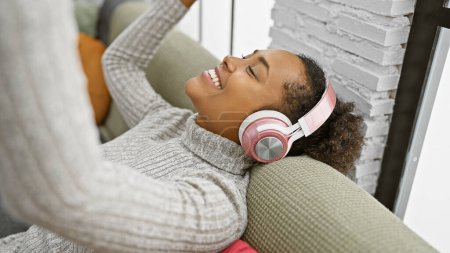 Téléchargez les photos : Une jeune femme souriante aux cheveux bouclés aime la musique sur écouteurs, allongée sur un canapé à l'intérieur. - en image libre de droit