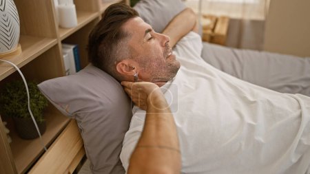 Foto de Hombre hispano joven agotado, un retrato de preocupación, acostado en su cama sufriendo de dolor cervical agonizante, descansando su cuerpo estresado sobre una almohada debajo de la manta, en un dormitorio débilmente iluminado - Imagen libre de derechos