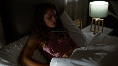 Téléchargez les photos : Une jeune femme réfléchie s'assoit au lit avec une lampe allumée dans une chambre faiblement éclairée, donnant une humeur contemplative. - en image libre de droit