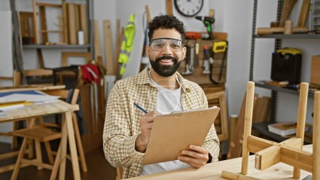 Téléchargez les photos : Un bel homme hispanique avec une barbe prend des notes dans un atelier de menuiserie, portant des lunettes de sécurité - en image libre de droit