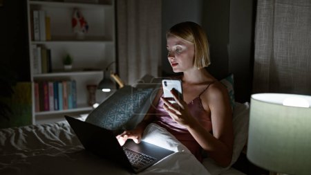Téléchargez les photos : Jeune femme blonde utilisant un smartphone et un ordinateur portable assis sur le lit dans la chambre - en image libre de droit