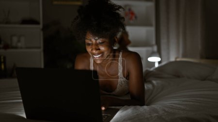 Téléchargez les photos : Femme afro-américaine portant de la lingerie à l'aide d'un ordinateur portable couché sur le lit dans la chambre - en image libre de droit