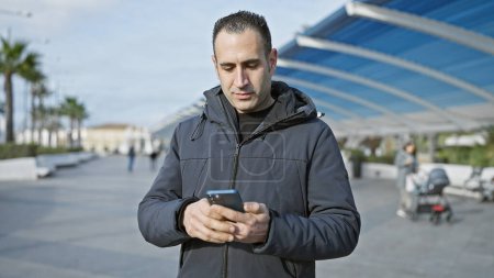 Photo for A young hispanic man in a jacket uses a smartphone while standing outdoors on a city promenade. - Royalty Free Image
