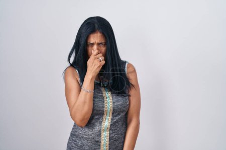 Téléchargez les photos : Femme hispanique mature debout sur fond blanc sentant quelque chose de puant et dégoûtant, odeur intolérable, retenant souffle avec les doigts sur le nez. mauvaise odeur - en image libre de droit