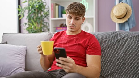 Foto de Hombre hispano joven guapo relajándose en casa, concentrado en mensajes de texto mientras bebe café en el sofá - Imagen libre de derechos