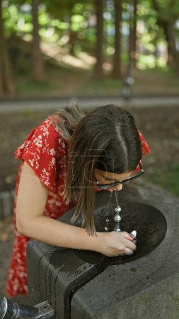 Téléchargez les photos : Superbe femme hispanique apaisant la soif avec une boisson rafraîchissante de la fontaine du parc, ses lunettes scintillant avec les éclaboussures de l'été ! beau portrait extérieur affirmant des choix sains et hydratants. - en image libre de droit