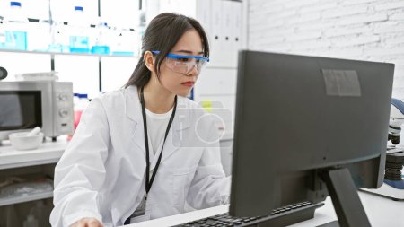 Photo for A focused young asian woman in a lab coat and safety glasses working on a computer in a modern laboratory. - Royalty Free Image