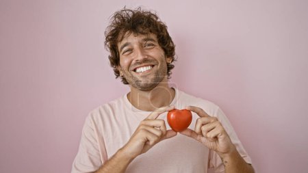 Photo for Smiling young hispanic man holding a red heart against a pink background portraying love and happiness. - Royalty Free Image