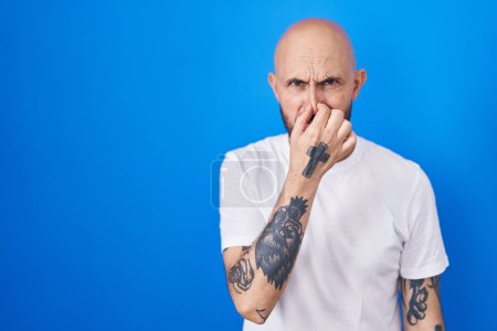 Téléchargez les photos : Homme hispanique avec des tatouages debout sur fond bleu sentant quelque chose de puant et dégoûtant, odeur intolérable, retenant souffle avec les doigts sur le nez. mauvaise odeur - en image libre de droit