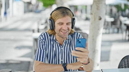 Téléchargez les photos : Un homme heureux avec casque bénéficie d'une terrasse ensoleillée tout en utilisant son smartphone dans un cadre urbain café rue. - en image libre de droit