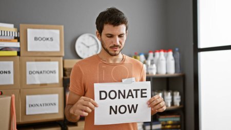 Hispanic man holding a 'donate now' sign in a warehouse filled with donation boxes.