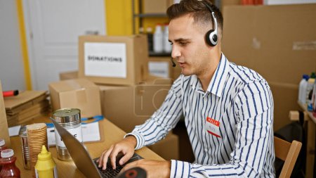 Foto de Joven guapo con auriculares trabajando en un ordenador portátil en una oficina de almacén rodeado de cajas y suministros de alimentos. - Imagen libre de derechos