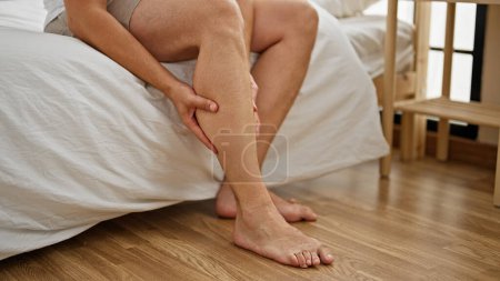 Téléchargez les photos : Homme aux cheveux gris assis sur le lit massant la jambe dans la chambre - en image libre de droit