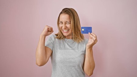 Téléchargez les photos : Thrilled jeune femme blonde célébrant avec confiance la victoire financière, tenant la carte de crédit avec un sourire joyeux sur fond rose isolé - en image libre de droit
