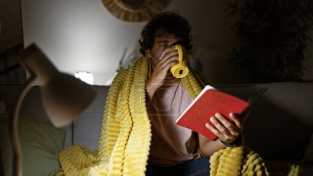 Foto de Hombre hispano guapo con barba y pelo rizado en casa sosteniendo un cuaderno rojo y una taza amarilla, envuelto en una manta amarilla en un sofá. - Imagen libre de derechos