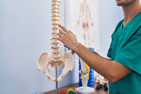Téléchargez les photos : Jeune homme caucasien physiothérapeute touchant le modèle anatomique de la colonne vertébrale à la clinique de réadaptation - en image libre de droit