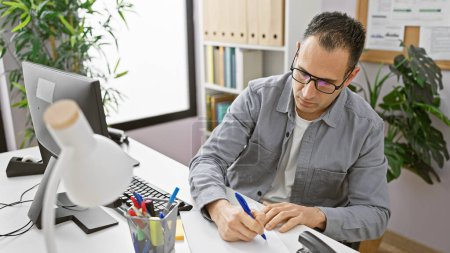 Téléchargez les photos : Homme hispanique concentré écrivant au bureau dans un bureau moderne lumineux, dépeignant le professionnalisme et la concentration. - en image libre de droit