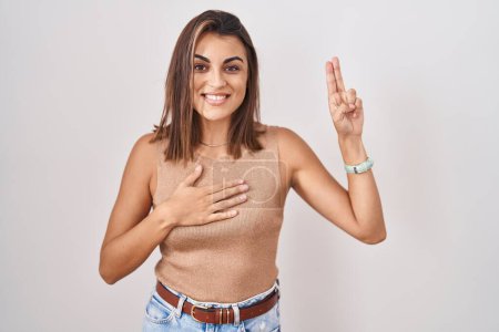 Foto de Mujer hispana joven de pie sobre fondo blanco sonriendo jurando con la mano en el pecho y los dedos hacia arriba, haciendo una promesa de lealtad juramento - Imagen libre de derechos