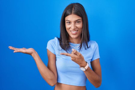 Photo for Brunette young woman standing over blue background amazed and smiling to the camera while presenting with hand and pointing with finger. - Royalty Free Image