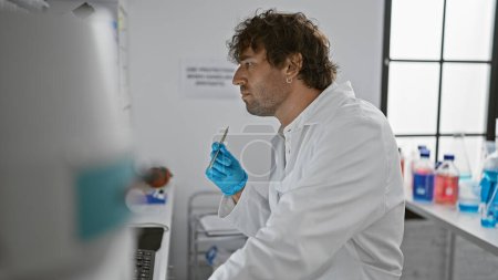 Photo for A focused man with a beard wearing gloves and a lab coat examines scientific equipment in a bright laboratory. - Royalty Free Image