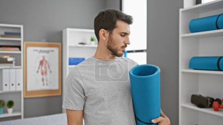 Photo for A young hispanic man with a beard holds a yoga mat in a physiotherapy clinic's modern, well-equipped interior. - Royalty Free Image