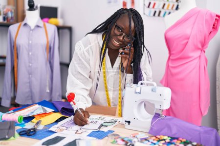 Téléchargez les photos : Femme afro-américaine tailleur parler sur smartphone dessin conception de vêtements au magasin de tailleur - en image libre de droit