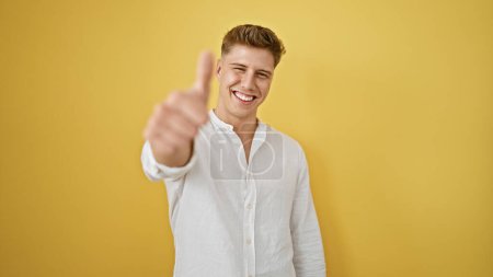 Photo for Happy young caucasian man, confidently standing, flashing a huge, joyful smile and giving an 'ok' thumb up sign, against a cheerful yellow isolated background. - Royalty Free Image