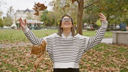 Foto de Una joven alegre lanza juguetonamente hojas de otoño en el aire en un entorno tranquilo parque, ejemplificando el ocio al aire libre. - Imagen libre de derechos
