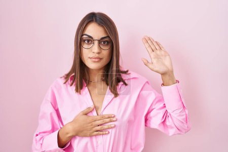Photo for Young hispanic woman wearing glasses standing over pink background swearing with hand on chest and open palm, making a loyalty promise oath - Royalty Free Image