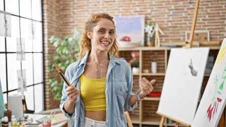 Photo for A cheerful young woman artist in a bright studio holding a paintbrush with a canvas on an easel and art supplies around. - Royalty Free Image